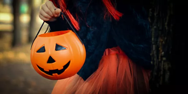 Happy girl wearing a witch's hat, with a pumpkin handbag, peeps out from behind a tree.