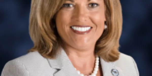 Portrait photo of a smiling white woman with blonde hair, wearing a gray suit and a white pearl necklace.